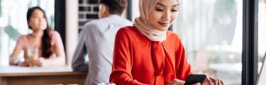 امرأة في قمة حمراء تجلس على مكتب باستخدام كمبيوتر محمول وهاتف محمول